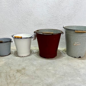 Op de afbeelding: Vier vintage emaille emmers in verschillende maten en kleuren. De emmers zijn grijs, wit, rood en grijs met een blauwe rand. Ze hebben houten handvatten.