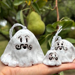 Peut inclure: Trois ornements fantômes en céramique blanche avec des traits noirs et une moustache. Chaque fantôme a une ficelle blanche pour être suspendu. Les ornements sont tenus dans une main, avec une poire et un feuillage vert en arrière-plan.
