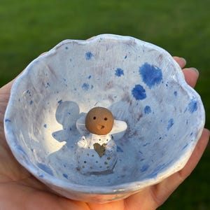Op de afbeelding: Een handgemaakte keramische kom met een lichtblauw en wit gespikkeld ontwerp. Binnenin een klein engelenbeeldje met een bruin hoofd, witte vleugels en een gouden hart. De kom wordt in een hand gehouden.