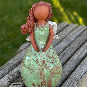 May include: A ceramic angel figurine with brown hair, a green dress with a tree design, and white wings.