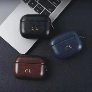 May include: Three leather AirPod cases in navy blue, black, and burgundy. Each case has the initials "C.L" in gold lettering. The cases are on a dark surface near a laptop.