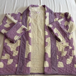 May include: A quilted jacket featuring a lavender and cream geometric pattern of semi-circles. The garment has a solid lavender trim along the front opening and cuffs, with a cream-colored interior lining and a visible tag at the inner neckline.