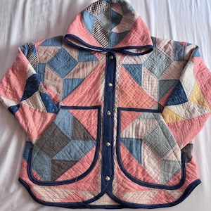 May include: A hooded jacket constructed from patchwork quilt fabric featuring a geometric pattern in shades of pink, blue, white, and brown. The garment includes a dark blue trim along the front opening, hood, and two large patch pockets with silver snaps.