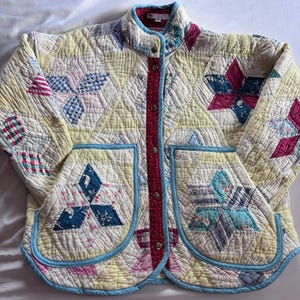 May include: A quilted jacket featuring a patchwork design of yellow, blue, and pink star patterns. The garment has a red button-down placket with white dots, two large front pockets with light blue trim, and a high collar with a quilted texture.