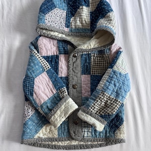 May include: A hooded jacket constructed from a patchwork of blue, white, and light pink fabric squares. The garment features a quilted texture, long sleeves with folded cuffs, and a front closure secured with three metal snaps and black trim.