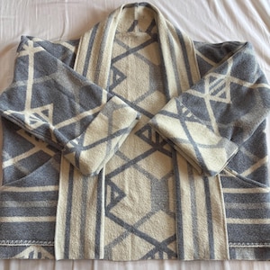 May include: A gray and cream-colored patterned cardigan. The open-front cardigan features a geometric design with gray and cream-colored shapes. The sleeves are long, and the cardigan has two front pockets.
