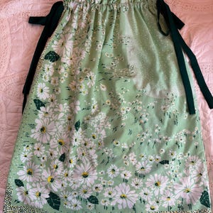 Puede incluir: Falda verde claro con un estampado floral de margaritas blancas. La falda tiene cintura con cordón y lazos verde oscuro. El borde inferior presenta un borde punteado. La tela es suave y ligera.