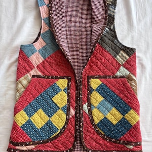 May include: A sleeveless quilted vest featuring a patchwork design with red, yellow, blue, and brown fabric squares. The garment has a brown floral trim, two front pockets, and a brown and white gingham lining with a small label at the neck.