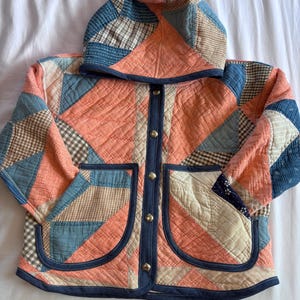 May include: A hooded jacket constructed from a patchwork of quilted fabric in shades of coral, blue, and beige. The garment features a front button closure with metallic snaps, two large patch pockets, and dark blue trim along the edges.