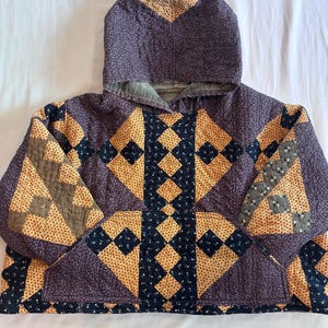 May include: A hooded pullover made from quilted fabric featuring a patchwork design. The garment displays a repeating pattern of dark purple, mustard yellow, and black floral and geometric prints arranged in diamond and square shapes with a front pocket.