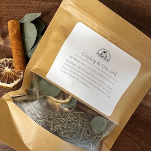 May include: A brown kraft paper pouch containing dried potpourri ingredients including rosemary, eucalyptus, and dried lemon slices. The label reads Unplug & Unwind and lists ingredients and instructions for simmering on a stove to release aroma.