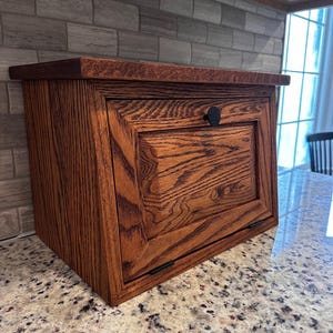 May include: A wooden bread box with a dark stain finish. The box has a hinged door with a black knob and a beveled top. The wood grain is visible, adding texture to the design. The bread box is sitting on a granite countertop.