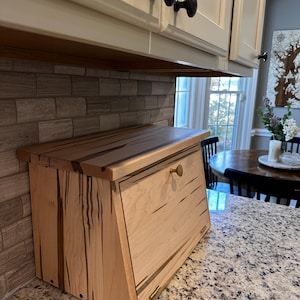 May include: A wooden bread box with a light-colored finish and dark wood grain accents. The box has a slanted front door with a brass knob and is hinged at the bottom. The lid is flat and has a similar wood grain pattern. The bread box is sitting on a granite countertop.