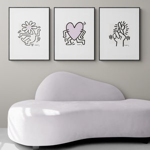 May include: Three black framed prints with white backgrounds and black line drawings of a person holding a flower, a person holding a heart, and a person holding up two fingers. The prints are hanging on a white wall above a white couch.