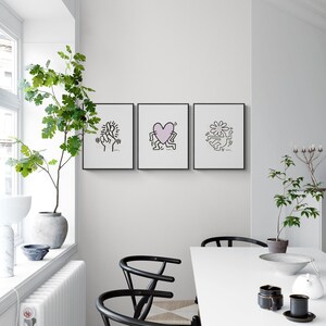 May include: Three black framed prints with white outlines of figures and a pink heart on a white wall. The prints are hanging above a white table with two black chairs.