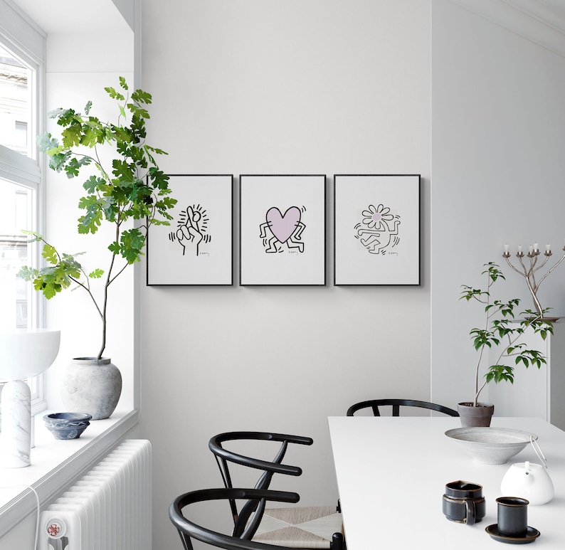 May include: Three black framed prints with white outlines of figures and a pink heart on a white wall. The prints are hanging above a white table with two black chairs.