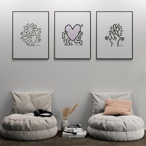 May include: Three black framed prints with white backgrounds and black line drawings of a flower, a heart, and a hand with crossed fingers. The prints are hanging on a white wall above two gray round poufs with cushions. A stack of books and a vase with dried flowers are on the floor in front of the poufs.