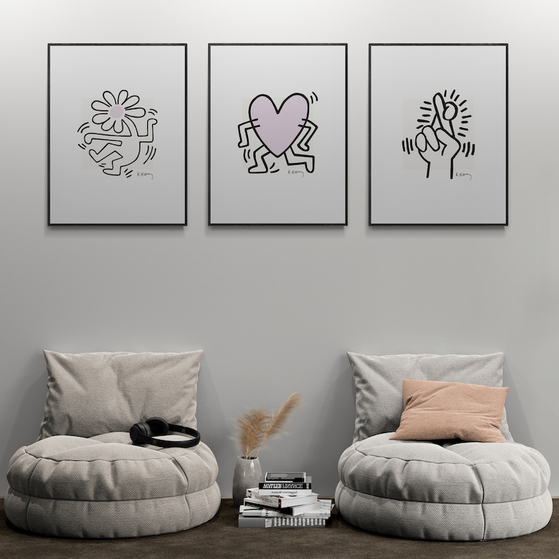 May include: Three black framed prints with white backgrounds and black line drawings of a flower, a heart, and a hand with crossed fingers. The prints are hanging on a white wall above two gray round poufs with cushions. A stack of books and a vase with dried flowers are on the floor in front of the poufs.
