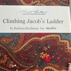 May include: A stack of fabric squares with a paisley design in burgundy, teal, and gold. The label reads "Climbing Jacob's Ladder" by Barbara Brackman for Moda.