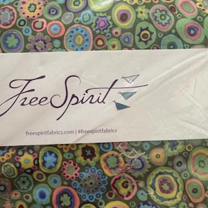 May include: Fabric with a colorful, abstract floral pattern in shades of green, blue, yellow, and purple. The fabric is packaged with a label that reads "Free Spirit" and "freespiritfabrics.com | #freespiritfabrics".