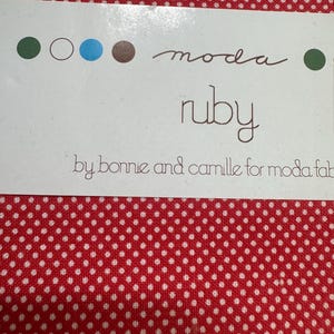 May include: Red fabric with a white polka dot design. A white label displays the text "moda ruby" and "by bonnie and camille for moda fab". The label also has four coloured circles: green, white, blue, and brown.