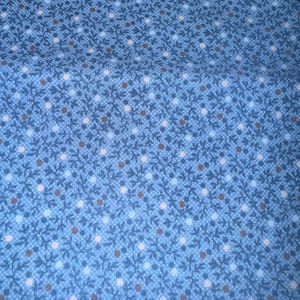 Puede incluir: Primer plano de una tela azul con un patrón repetitivo de pequeños puntos blancos y marrones, y elementos florales azul oscuro. La tela tiene una apariencia texturizada, con un tejido sutil en forma de rejilla visible en todo el diseño.