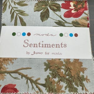 May include: A fabric bundle with floral patterns in red, green, and brown on a pale blue backdrop. The label reads "Sentiments" by 3 sisters for moda. The fabric is cut into squares, with the edges visible.