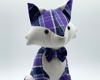 May include: A plush fox toy with a purple and black plaid pattern and a black bow tie. The toy is sitting on a white surface.