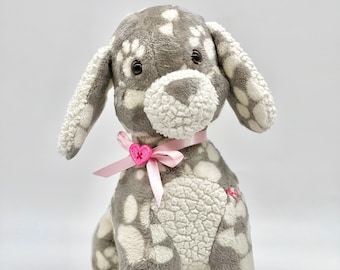 May include: A gray and white plush dog toy with paw print patterns. The dog has a pink ribbon around its neck with a pink heart button.