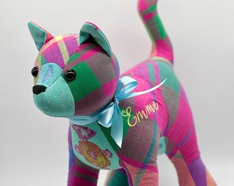 May include: A handmade stuffed cat in a standing pose, crafted from colorful plaid fabric. The cat features a teal face and ears, black eyes, and a blue ribbon with the name "Emme" embroidered on it. The cat's body is a mix of pink, green, and purple.