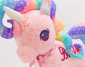 May include: A plush unicorn toy in pink fabric with a white swirl pattern. The unicorn has a rainbow mane and tail with white polka dots. The word "Bella" is embroidered on the side, and a pink ribbon is tied around its neck.