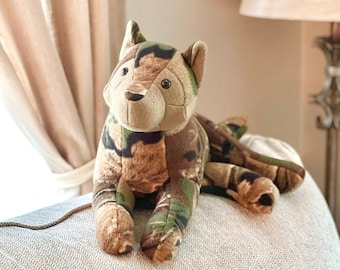 May include: A plush toy in the shape of a cat, featuring a camouflage pattern in shades of brown, green, and tan. The cat is lying down on a light-colored surface, with its head and paws visible. The toy has a soft, cuddly appearance.