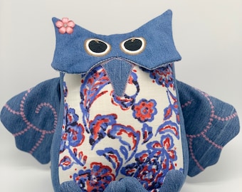 May include: A blue denim owl with a floral pattern on its chest. The owl has large black eyes and a pink flower on its head.