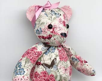 May include: A plush teddy bear with a floral print fabric. The bear is wearing a pink bow and has black eyes. The fabric features pink, blue, and green flowers and butterflies.