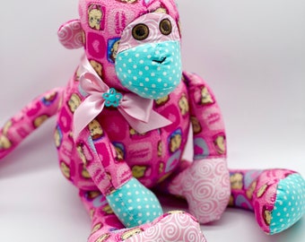 May include: A pink monkey plush toy with a blue and white polka dot face mask. The monkey is wearing a pink ribbon with a blue flower.