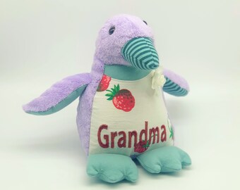 May include: Purple penguin plush toy with a white and green striped beak, a white flower, and the word "Grandma" embroidered on the chest. The penguin is wearing a white shirt with red strawberries.