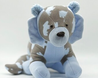 May include: A plush toy in the shape of a dog with a gray and white patterned body and blue ears and legs. The toy has a brown nose and black eyes.