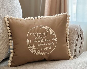 May include: A brown throw pillow with white pom-poms around the edge. The pillow has an embroidered design that says "In Memory of a life so beautifully lived, a heart so deeply Loved".