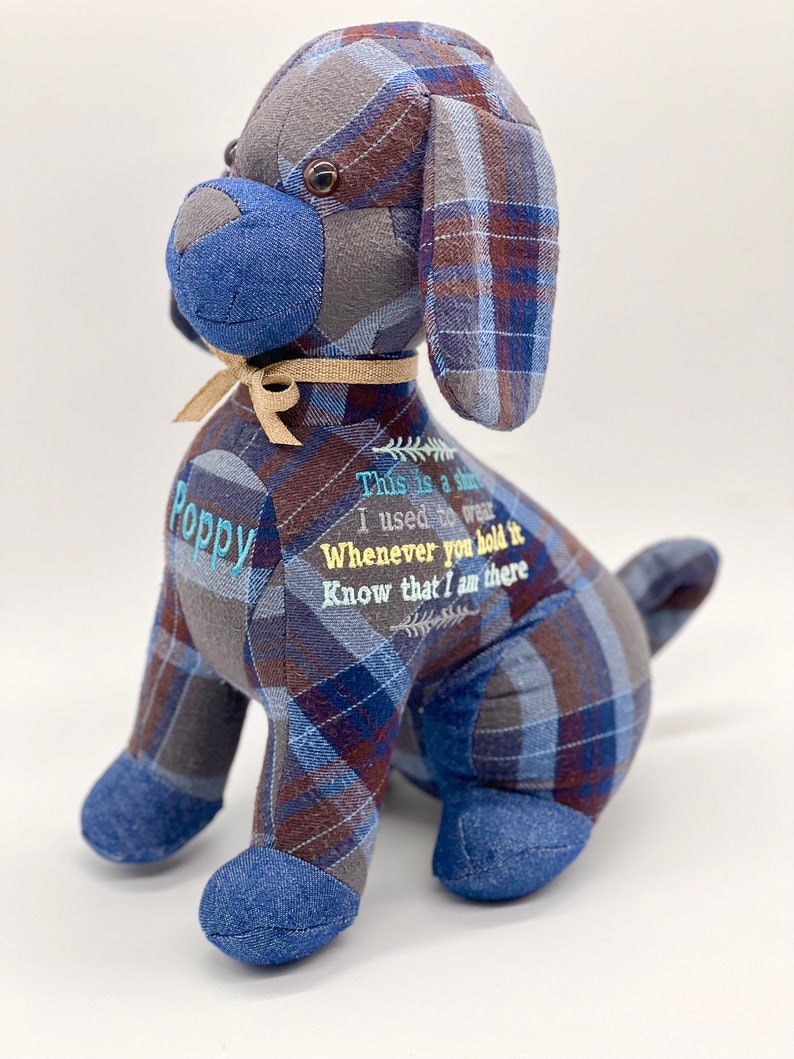 May include: A blue and gray plaid dog-shaped stuffed animal with a tan bow. The dog has a brown button eye and the text "This is a shirt I used to wear Whenever you hold it Know that I am there" is embroidered on the dog's chest. The dog's name "Poppy" is embroidered on the left side of the dog.