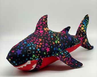 May include: A plush toy shark with a black and rainbow star pattern. The shark has a red mouth with white teeth and a black eye.