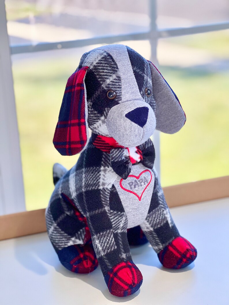 May include: A plush dog toy with a black and white plaid pattern, red and black plaid ears, and a gray face. The dog is wearing a red bow tie and has a red heart with the word "PAPA" embroidered on its chest.