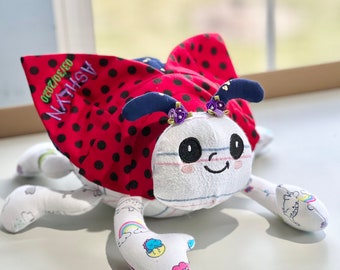 May include: A red and black ladybug plush toy with a white and blue striped body. The ladybug has a smiling face with black eyes and pink cheeks. The wings are decorated with black polka dots and the text "ASHLYN 09/20/2020" is embroidered on the left wing.