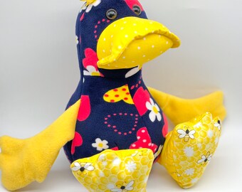 May include: A plush toy duck with a yellow beak and feet. The duck is wearing a blue and red fabric with a pattern of hearts and flowers. The duck has a yellow bow with a red strawberry on top of its head.