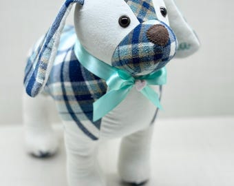 May include: A stuffed dog toy with a white body and blue plaid patterned head, ears, and back. The dog has a brown nose, black eyes, and a light blue ribbon tied in a bow around its neck. A small pink flower is attached to the bow.