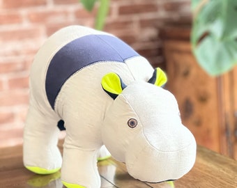 May include: A plush toy hippopotamus with a white body, gray and blue striped back, and yellow ears. The hippo is standing on a wooden surface.