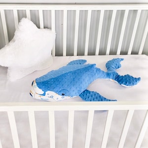 May include: A blue whale plush toy with white and orange accents lies in a white crib. The whale has a textured fabric and a friendly expression.