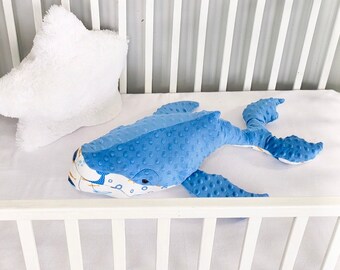 May include: A blue whale plush toy with white and orange accents lies in a white crib. The whale has a textured fabric and a friendly expression.