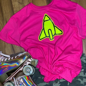 May include: A bright neon pink short-sleeve t-shirt featuring a large, lime green rocket ship graphic with a thick black outline, paired with camouflage print pants and a pair of metallic, iridescent roller skates with light-colored wheels.
