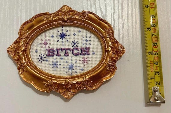 Hand-stitched sweary embroidery that turns grandma's craft into a delightfully rude wall statement — the contrast is the whole joke.