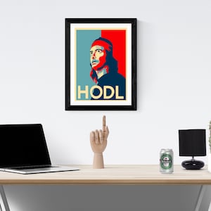 May include: A framed poster with a blue, red and white design. The poster features a man with long hair and a red hat. The text "HODL" is printed in yellow at the bottom of the poster. The poster is hanging on a white wall above a wooden desk.